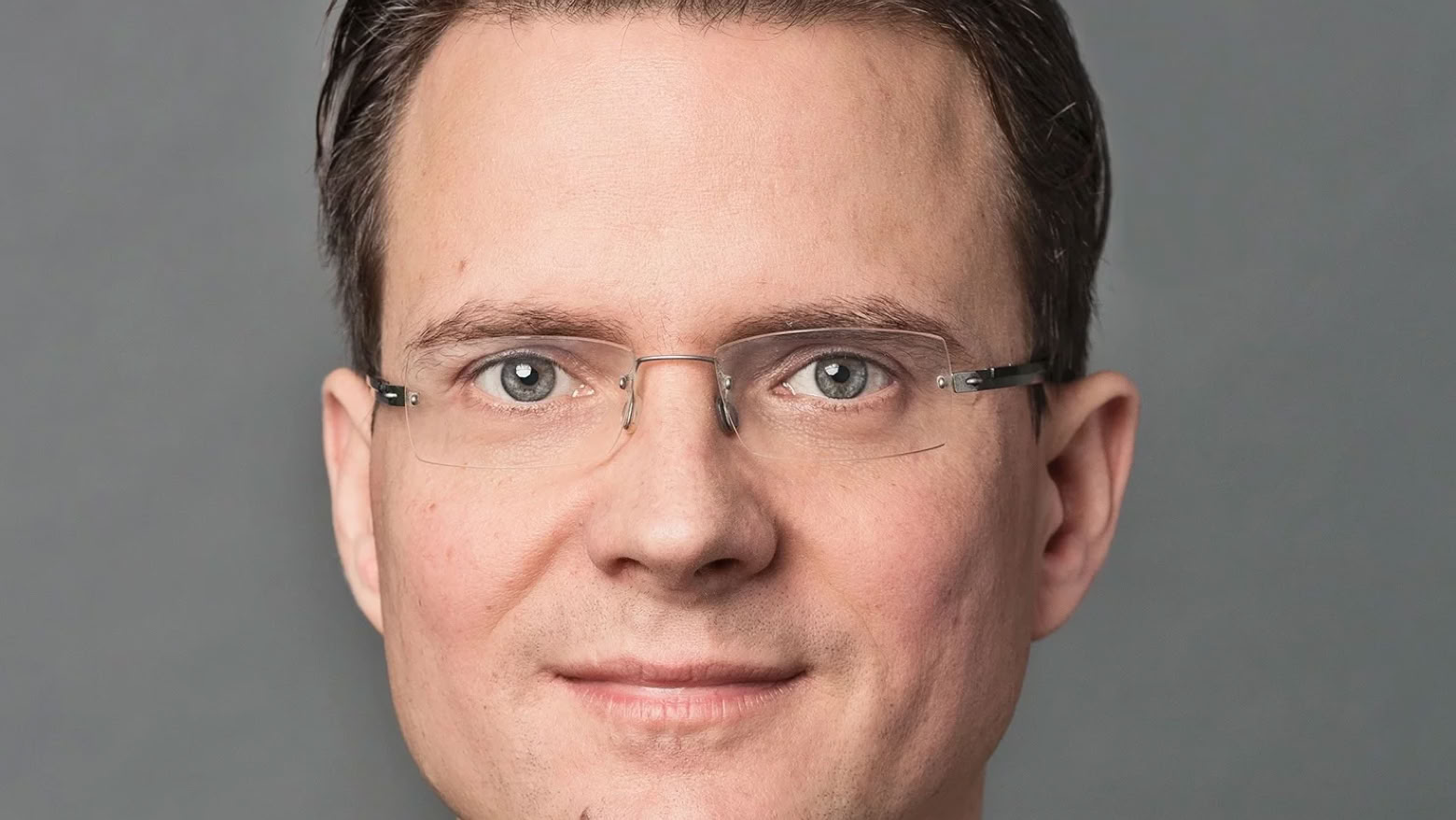 Mann mit Brille und Anzug vor grauem Hintergrund.