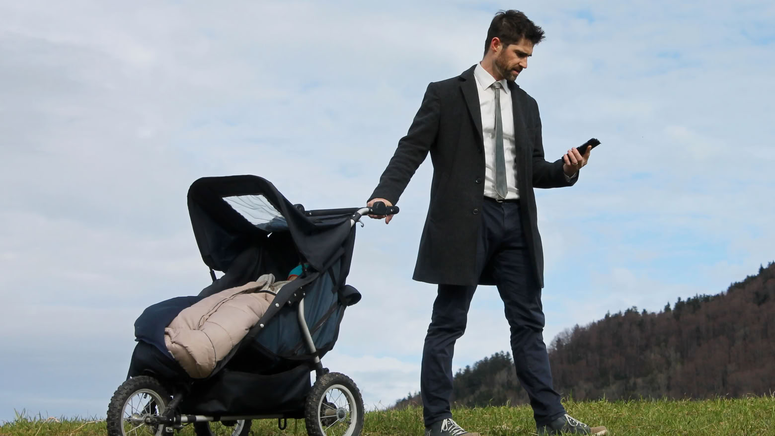 Ein Mann im Anzug steht auf einer Wiese und hält einen Kinderwagen mit einer Hand, während er mit der anderen Hand auf sein Smartphone schaut.