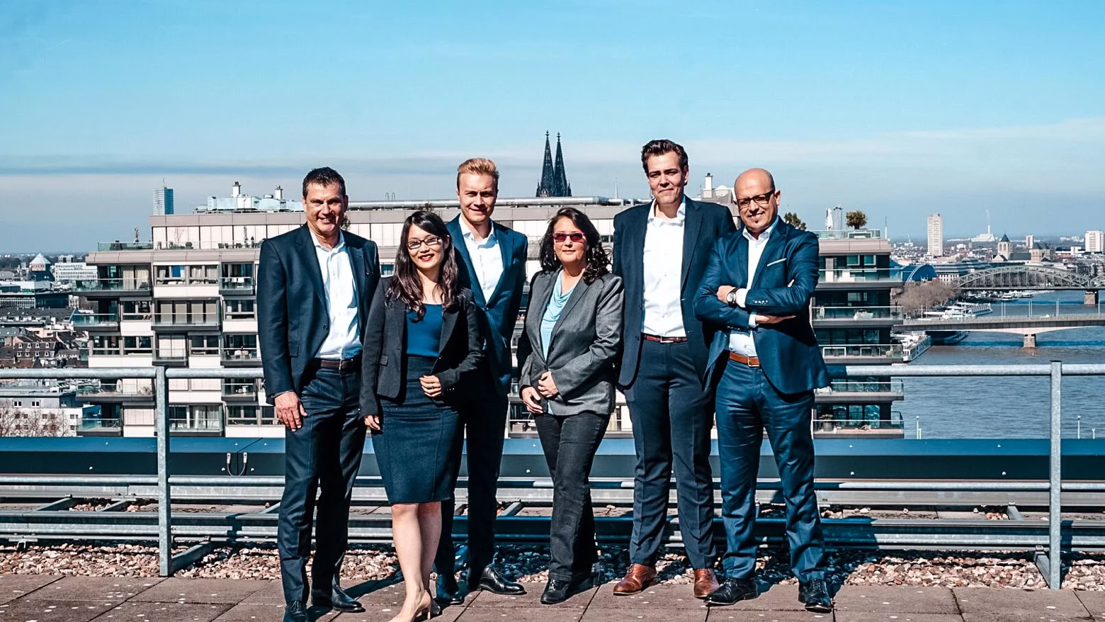 Sechs Personen in formeller Kleidung stehen auf einem Dach mit Stadtlandschaft im Hintergrund.